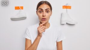 Waist up Shot of Shocked Brunette Woman Keeps Hand on Chin Stares at Camera Has Stupefied Expression Dressed in Casual Basic T Shirt Poses against White Background with Plastered Sneaker and Boot